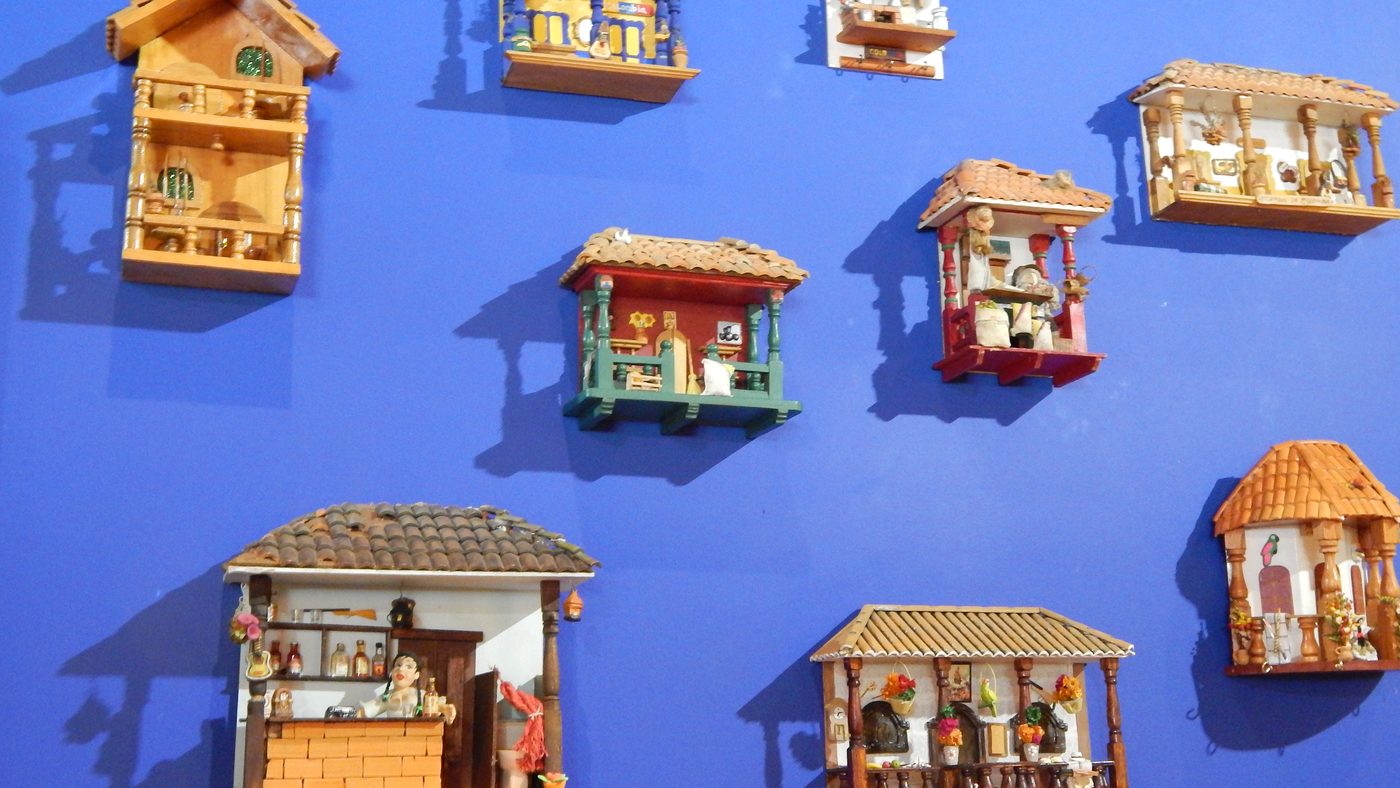 Nine miniature Colombian pueblito houses with terracotta tiled roofs and tiny balconies, mounted on a deep blue wall