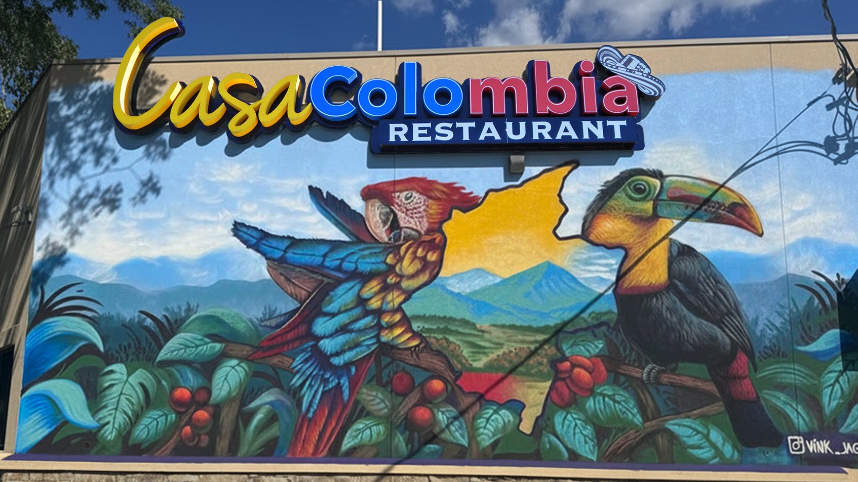 The Casa Colombia storefront on East 7th Street — the lit Casa Colombia Restaurant sign above a painted mural of a scarlet macaw and a keel-billed toucan perched among coffee branches, with the map of Colombia and the Andes behind them